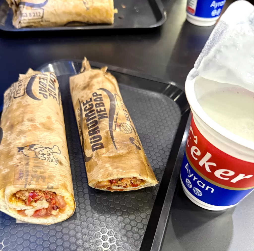 Adana kebab dürüm, mleté maso na špízu v placce, na podnosu. Ayran v kelímku.